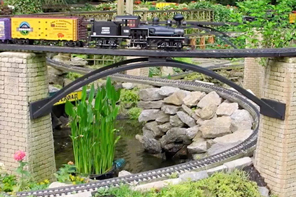a garden railway showing a steam locomotive on a bridge over a pond and rocks