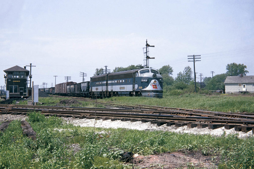 Silver and blue train rolling on track surrounded by grass under a cloudy sky.