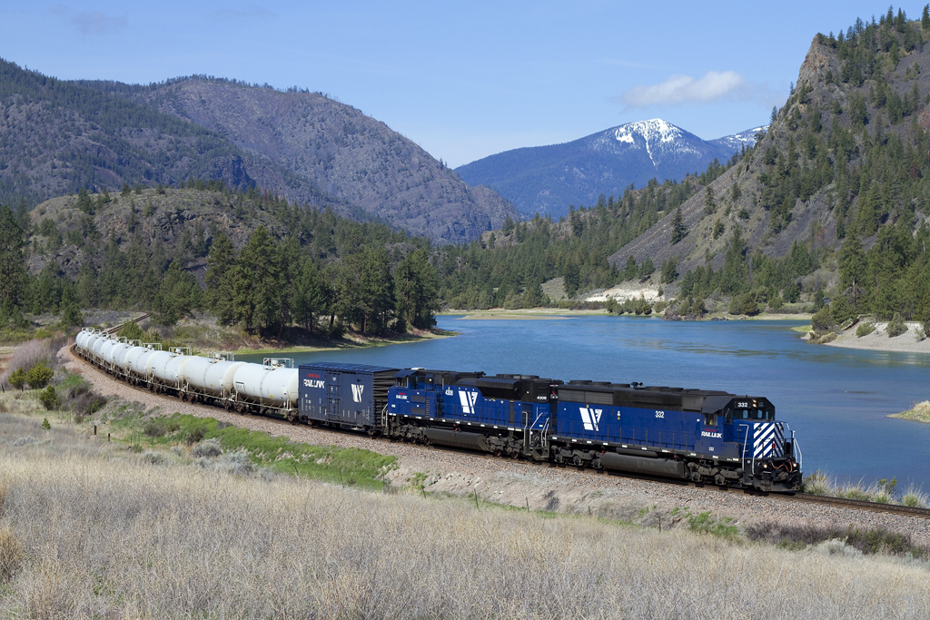 A blue train turning a corner by a body of water with mountains in the background