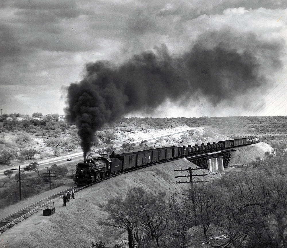 A black and white photo of locomotive Texas & Pacific 2-10-4 with smoke coming out of its chimney. People are standing off to the side of the tracks to let the train pass.