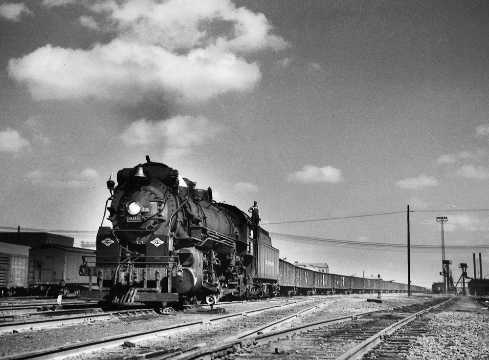 A black and white photo of a brakeman standing on top of locomotive Texas & Pacific 2-8-2 805 as it pulls out of the rail yard