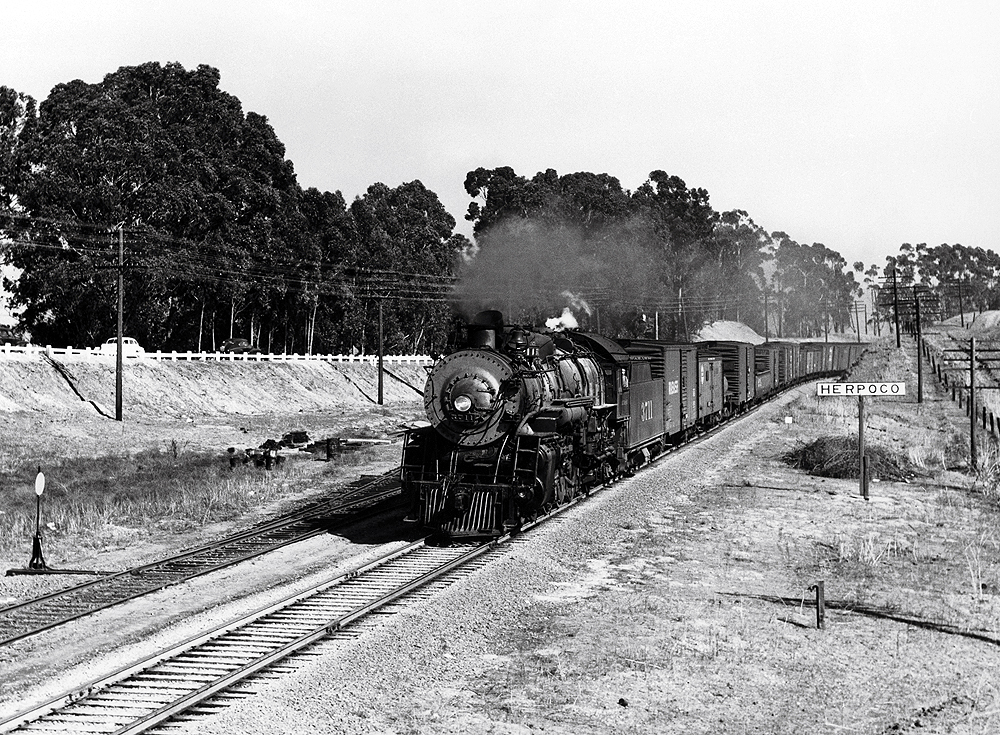 A black and white photo of locomotive 4-8-2 3711 on the tracks passing a Herpoco sign