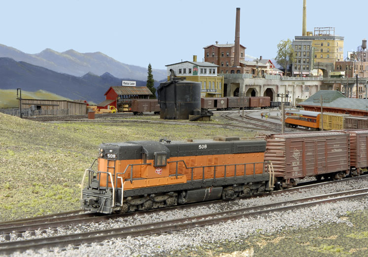 A black and orange model locomotive leads a train out of a city on the Milwaukee Road North Montana Line