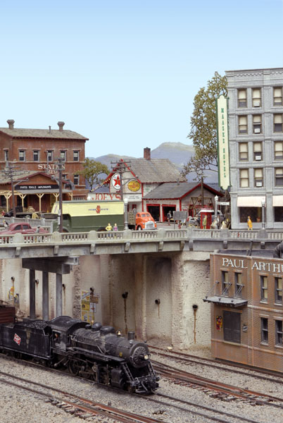 A black steam locomotive leads a train under a bridge in a city on a model railroad layout