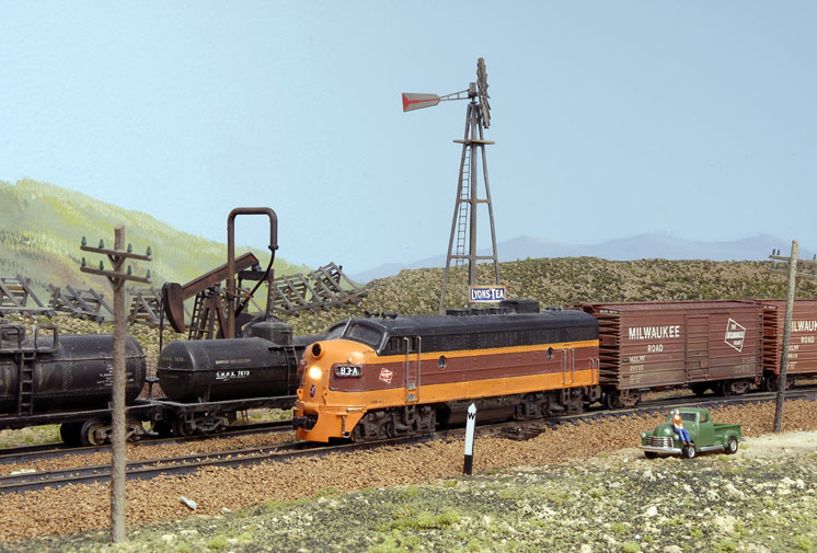 A black and orange streamlined locomotive leads a train past a parked green truck on the Milwaukee Road North Montana Line