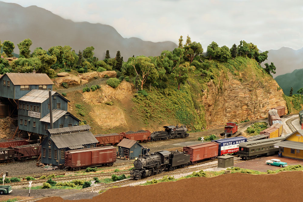 Image of a rural rail yard in a mountainous, arid, verdant landscape with multiple steam locomotives running trains.