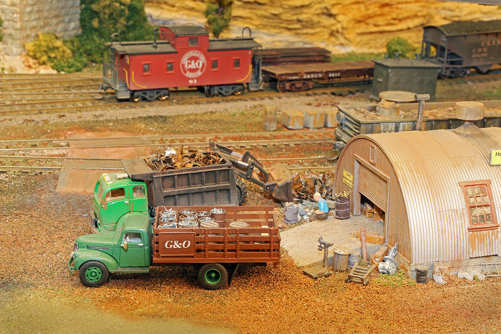 Model trucks loaded with scrap metal near a quonset hut in a rural rail yard.