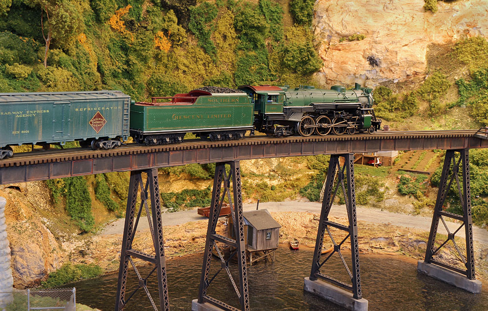 A green steam locomotive hauling a passenger or express train over a trestle in a verdant, arid landscape.