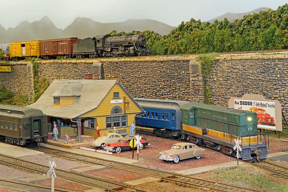 A diesel locomotive and a passenger car near a small town train station. A steam locomotive hauls a freight train on a higher level in the background.