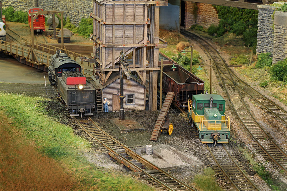 A steam and diesel locomotive operate near a coaling tower and a round table in a verdant rail yard scene.