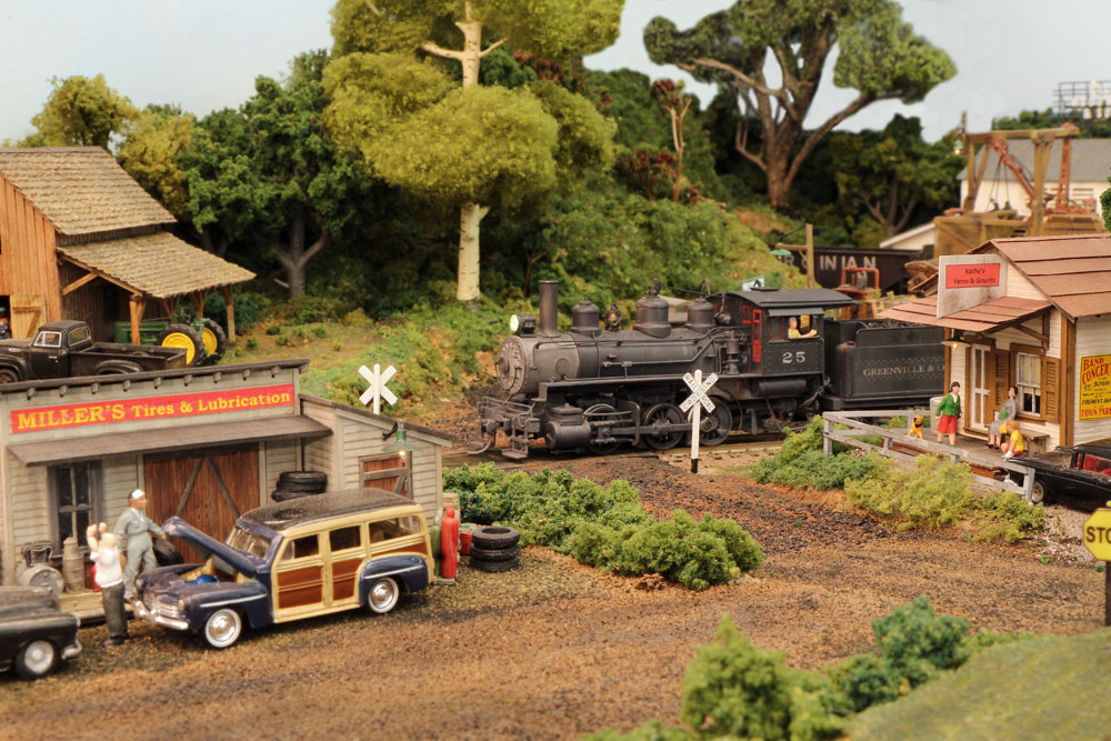 A steam locomotive switcher pulls a train through the outskirts of a small, rural town with dirt roads.