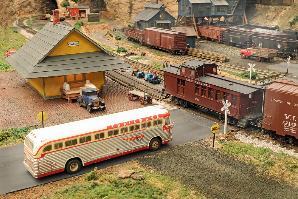 A passenger bus waits at a crossing for a caboose-tailed freight train next to a small town passenger station.