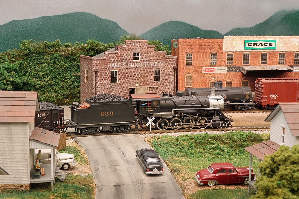 A 2-8-0 steam locomotive hauls a freight train through an industrial scene bracketed by 19th century brick buildings.