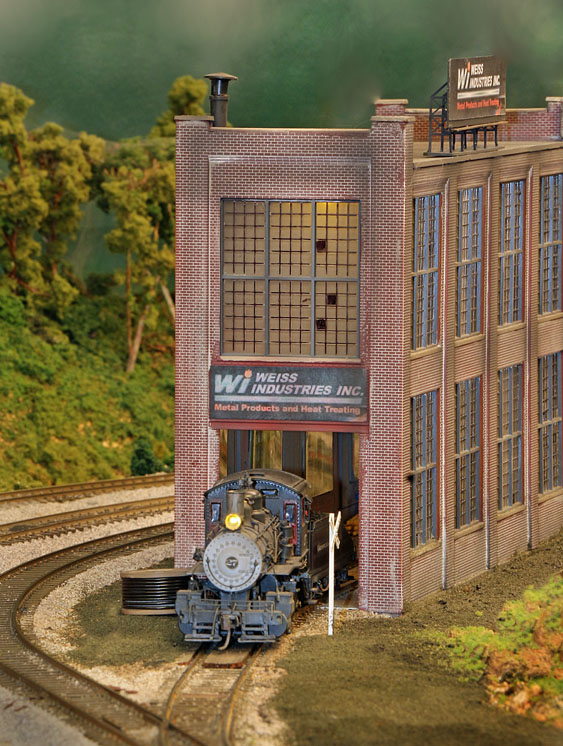 A steam locomotive pokes out of the corner railroad entrance of a multi-story brick building.