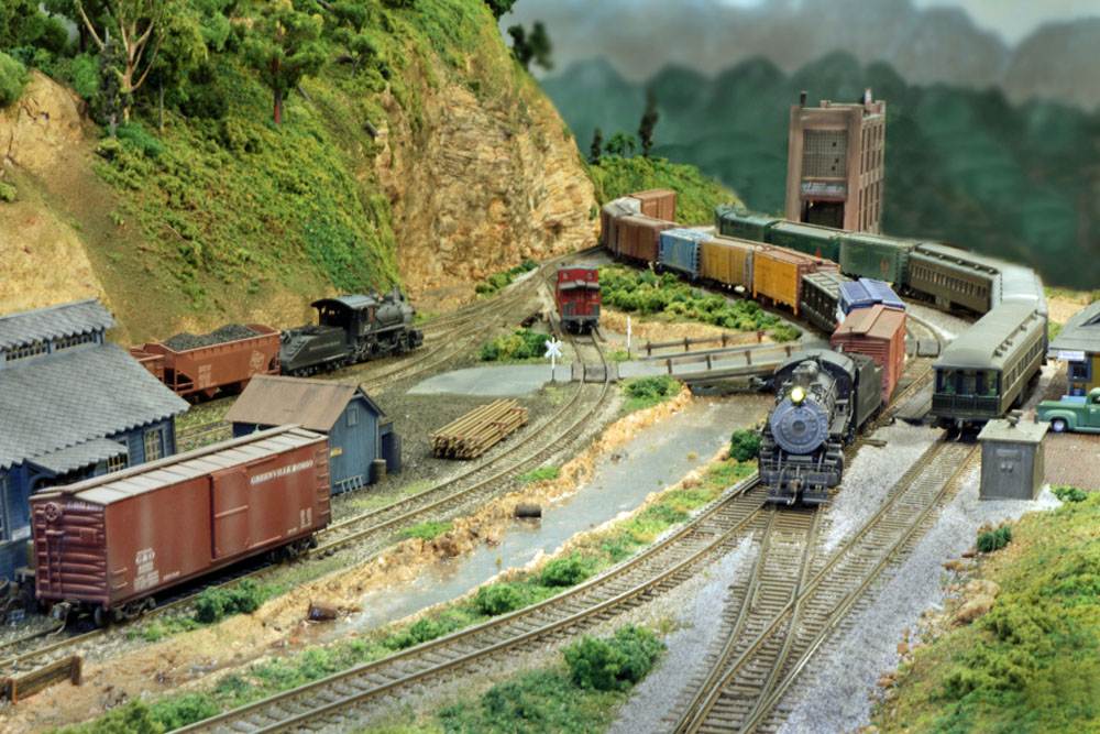 A steam locomotive hauls a freight train through a rural rail yard.
