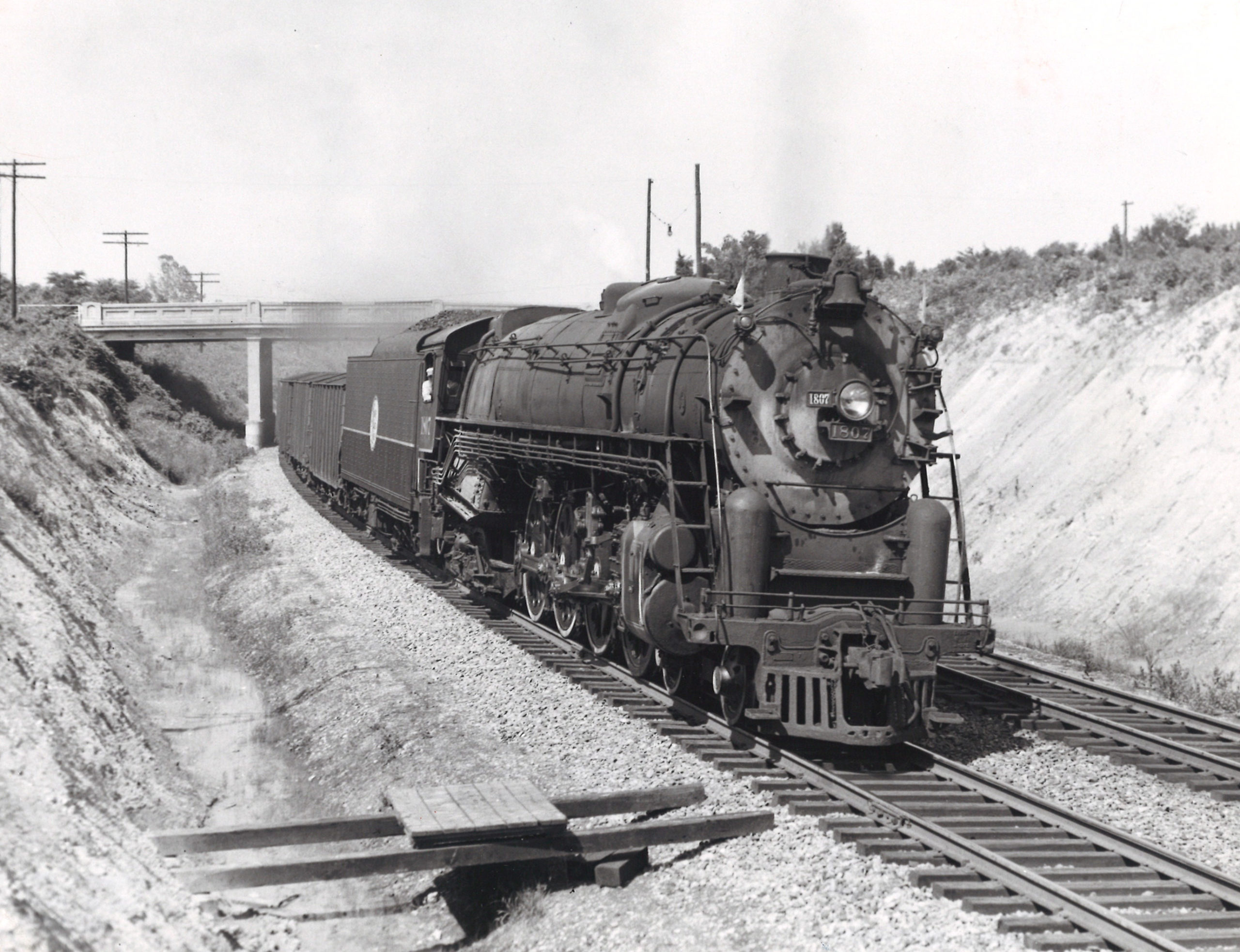 A black and white close up photo of a steam locomotive moving between two hills