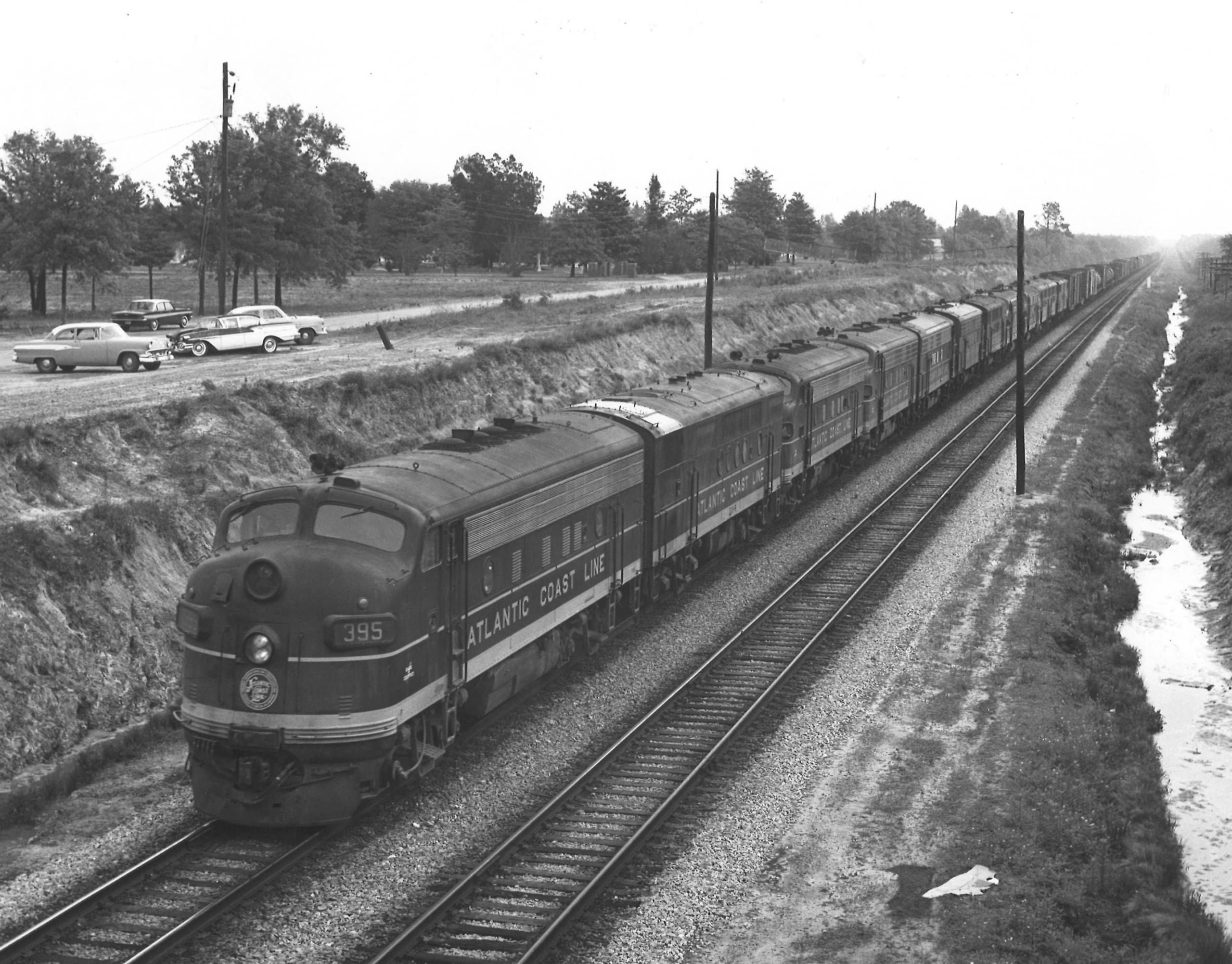A black and white diesel locomotive on the tracks next to a parking lot with cars in it