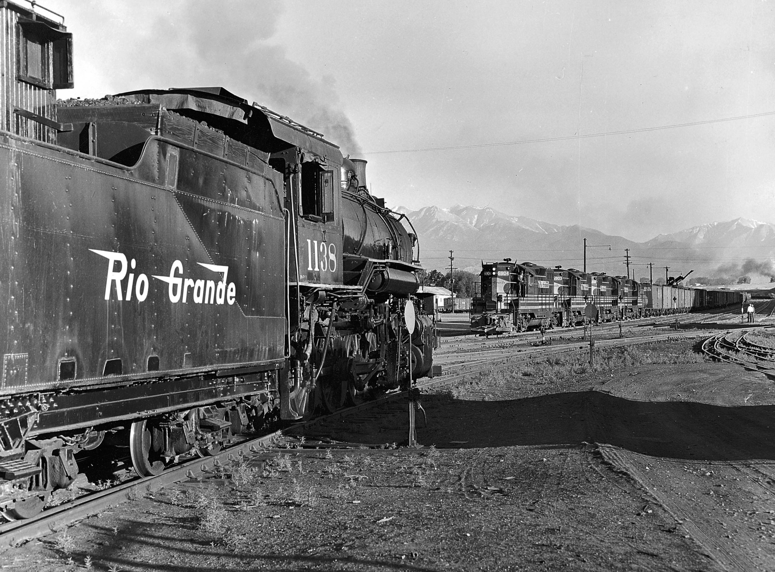 A black and white close up of a steam locomotive approaching a diesel locomotive on the tracks.