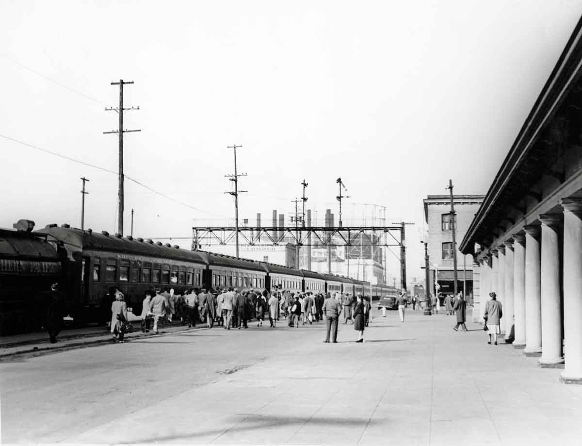 Crowd on platform to board passenger train with gas holder in distance