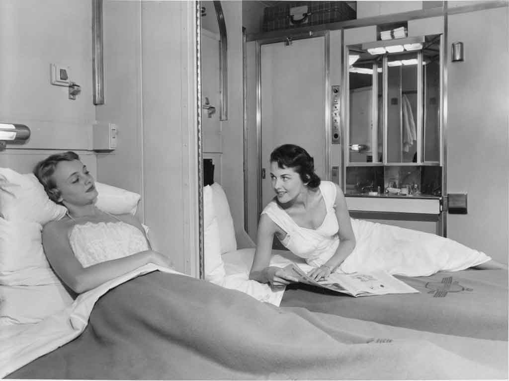 A couple of women laying in their beds talking through an open partition