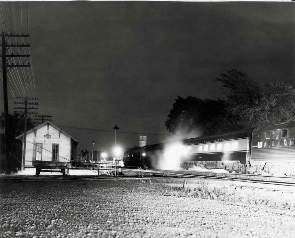 A black and white photo of a passenger train passing a station in the dark