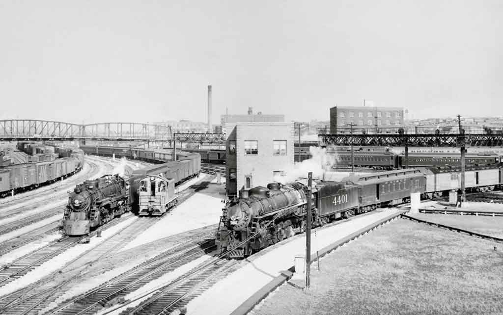 Two steam-powered passenger trains pass diesel switcher under bridges