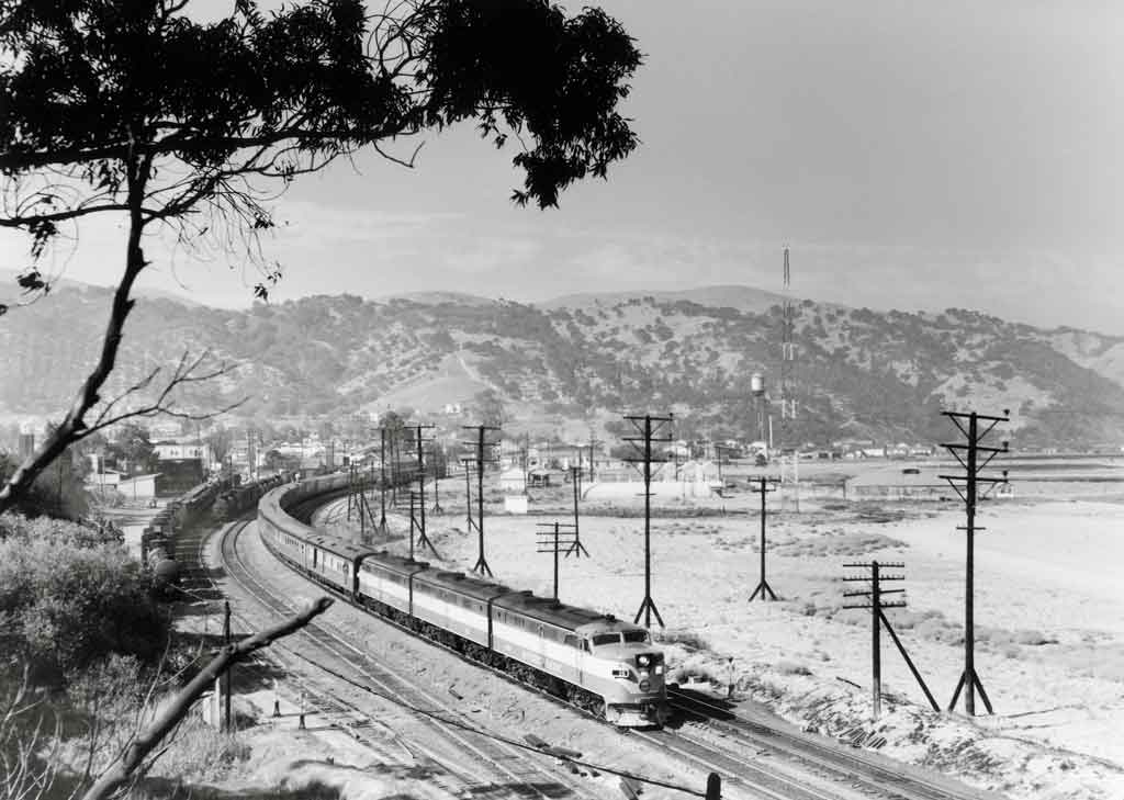 A black and white photo of a locomotive turning a corner with mountains in the background