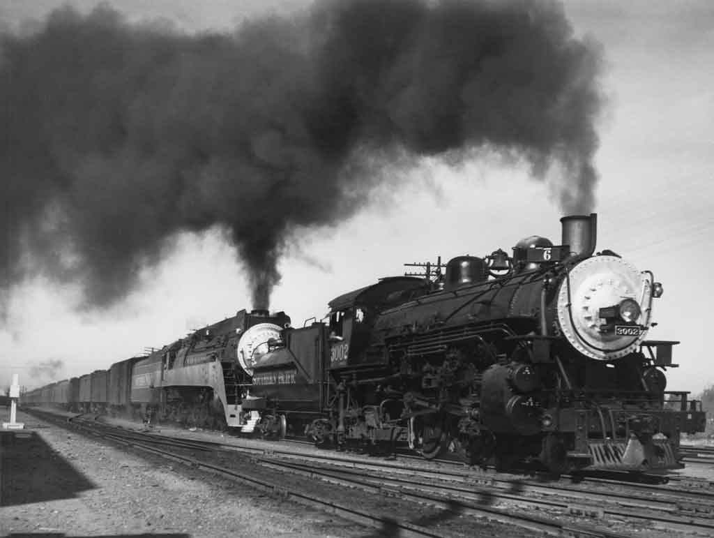 A black and white photo of two locomotives on the same track together with black smoke coming out of their chimneys