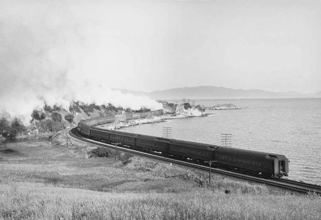 A black and white photo of a locomotive turning a corner next to a lake