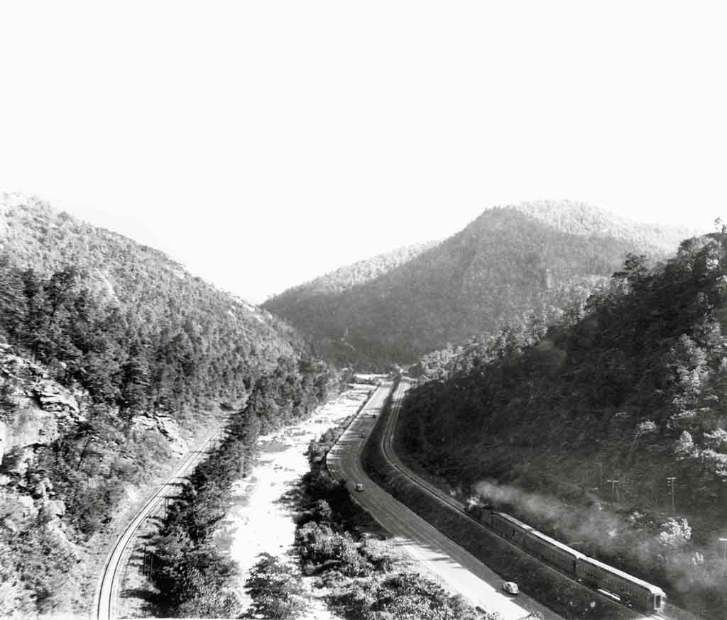A black and white birds eye view of a train moving through mountains 