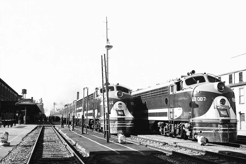 A black and white photo of two locomotives picking up passengers
