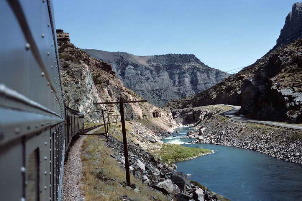Looking back at a train as it passes through a mountain and body of water