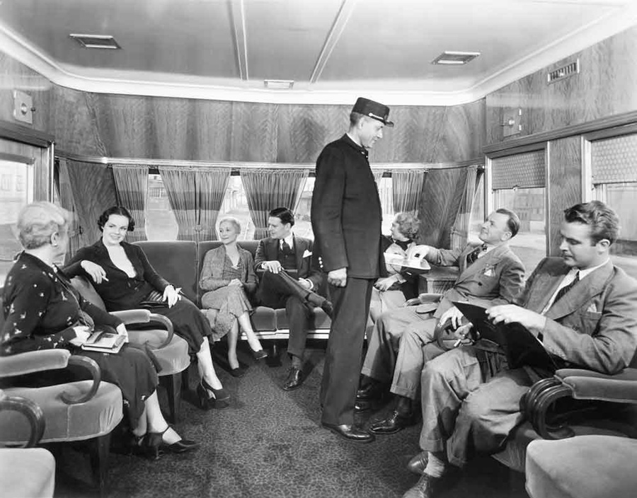 A black and white photo of a group of people sitting and talking while riding a train