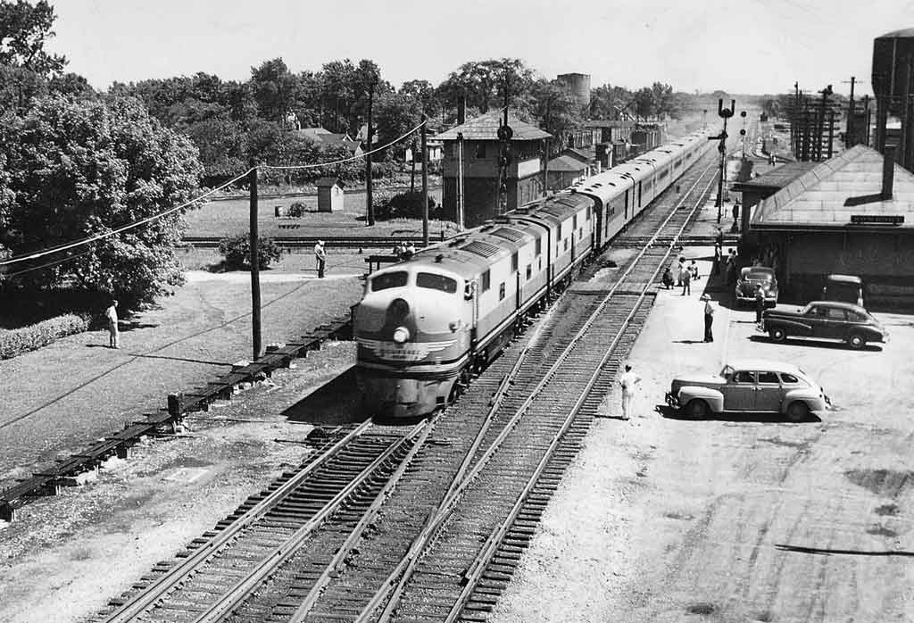 A black and white photo of a locomotive passing people and cars