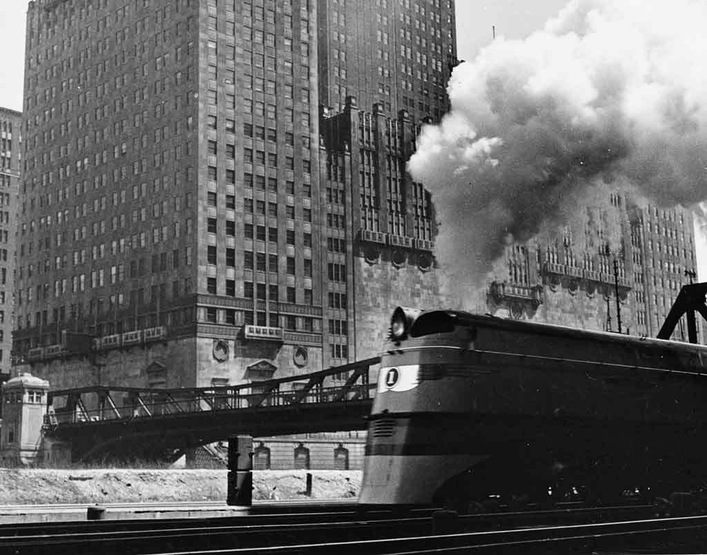 A black and white photo of a locomotive with white smoke coming out of its chimney while passing by a city