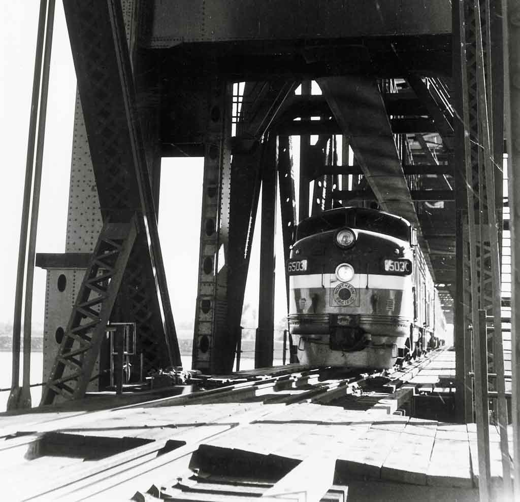 A close up black and white photo of a train coming through a bridge