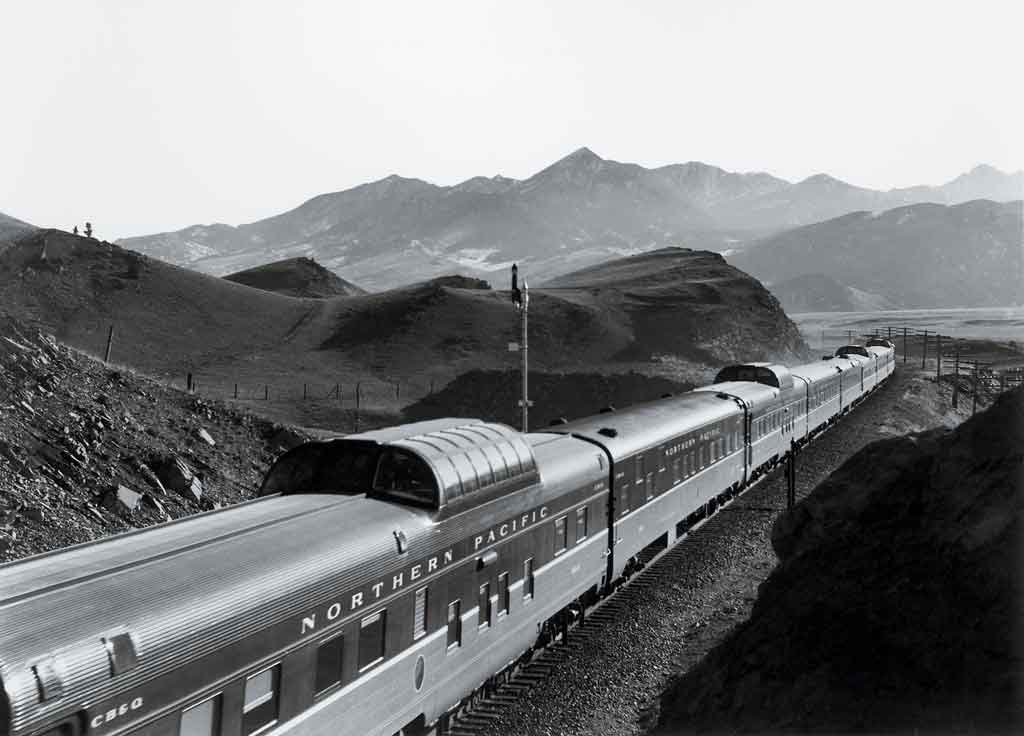 A black and white picture taken overhead a long locomotive moving through the mountains