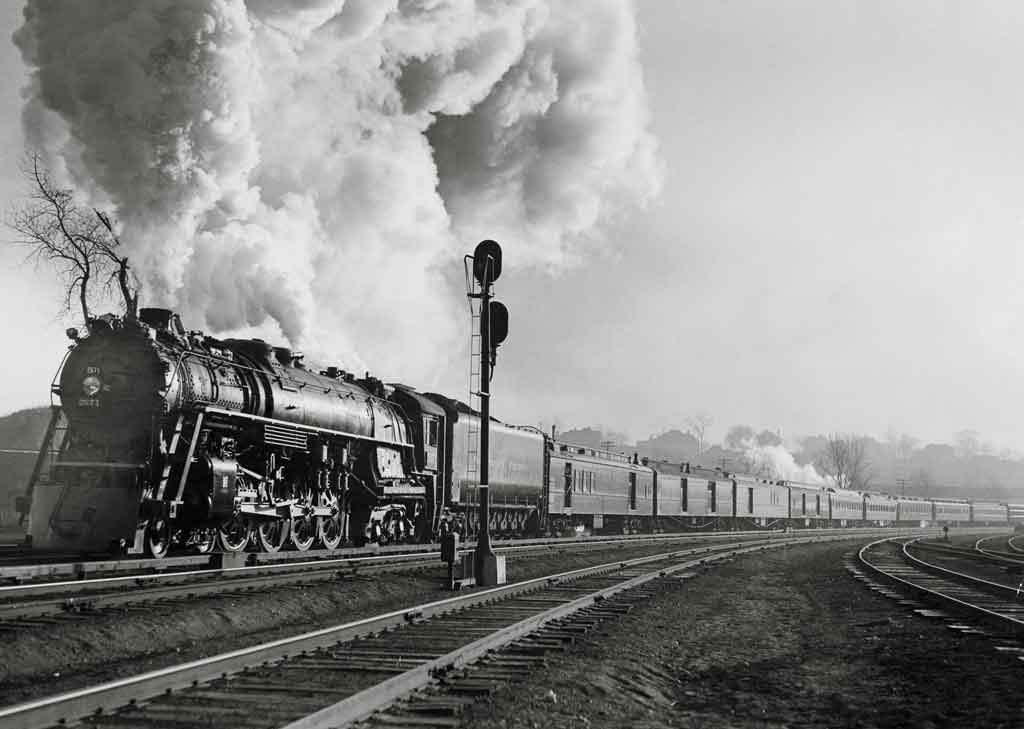 A black and white photo of a locomotive with big white smoke coming out of its chimney