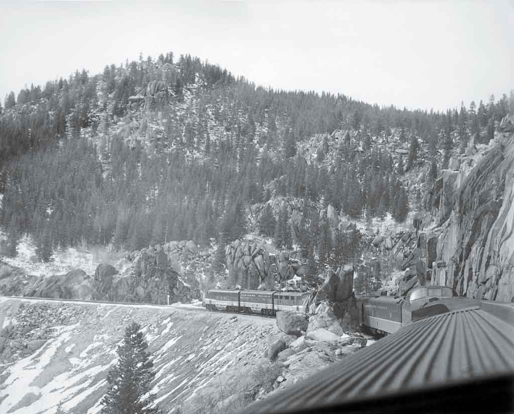 A black and white photo taken from the top of a locomotive looking back at the train and the mountains