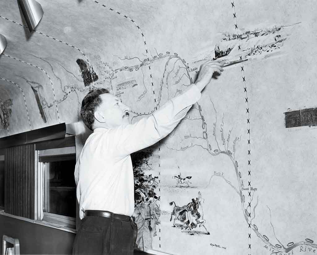 A black and white photo of an artist pointing at something on his mural inside a passenger car