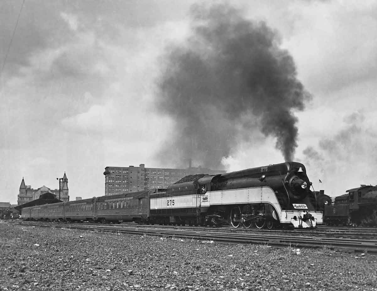 A black and white photo of a train with black smoke coming out of its chimney as it passes through a city
