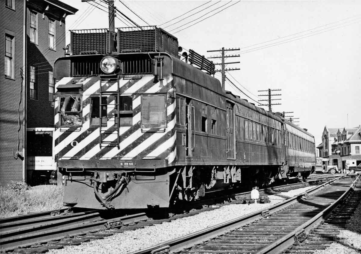 A black and white photo of a stiped locomotive passing by buildings
