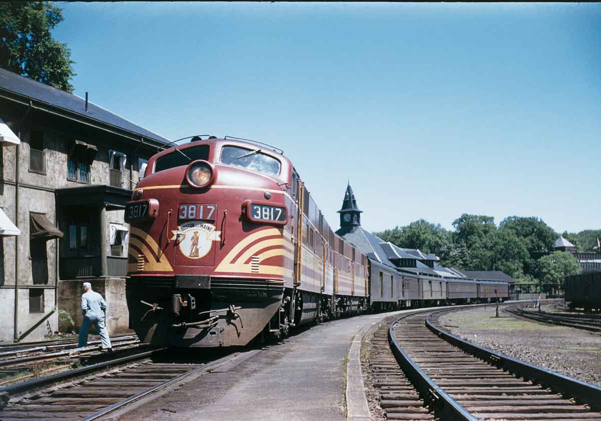 A yellow and red train passing through a town as a person crosses the tracks