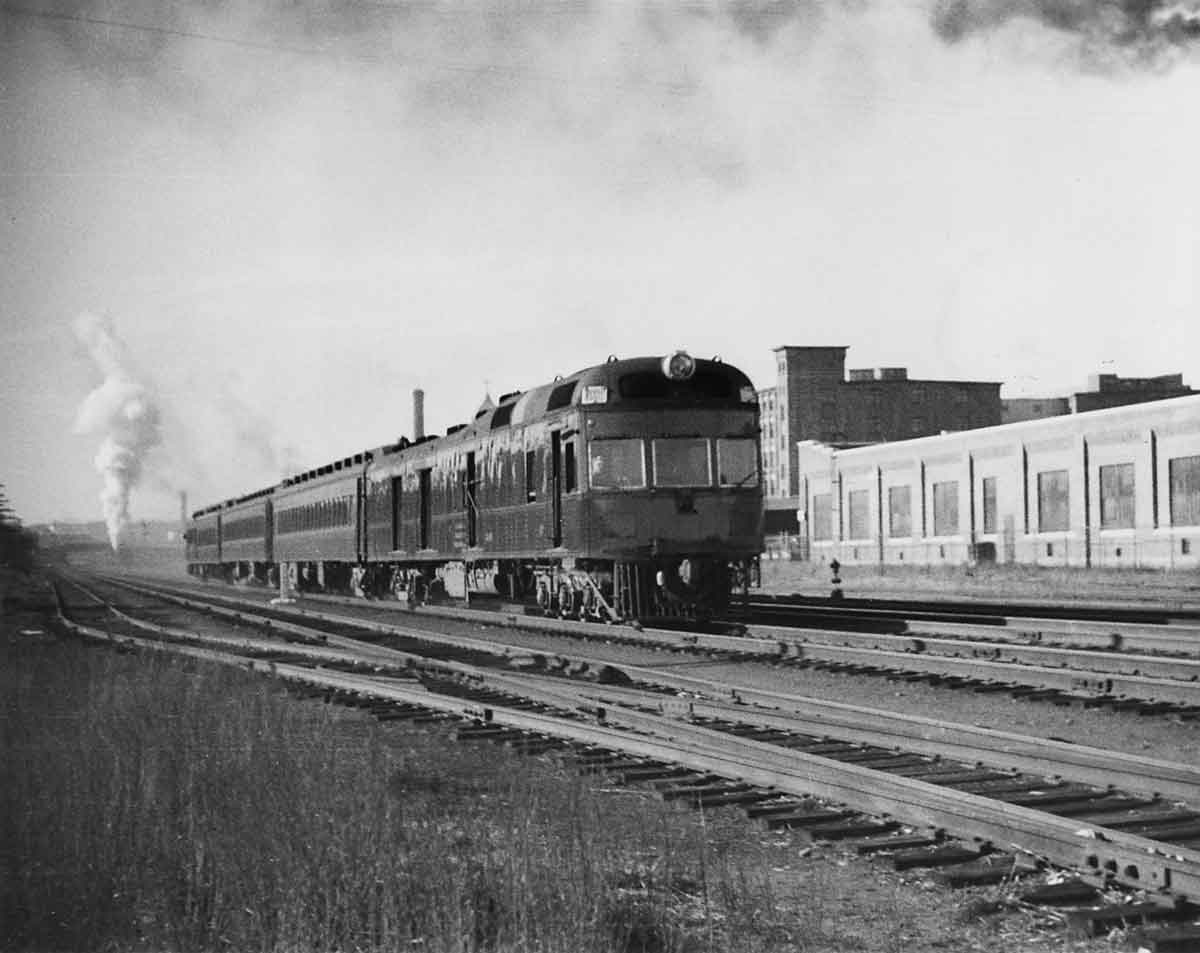 A black and white photo of a car baggage-RPO-locomotive passing buildings on the track