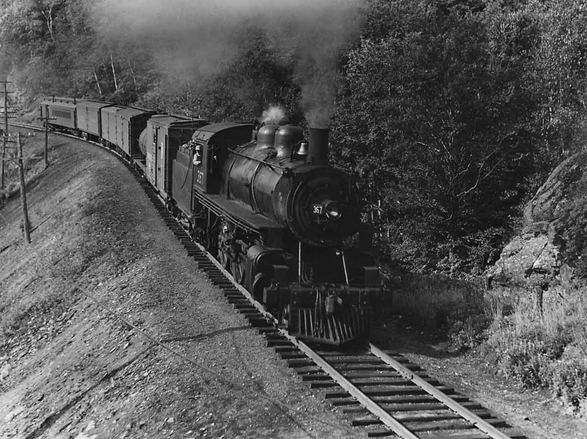 A black and white photo of a locomotive with smoke coming out of its chimney as it moves through the trees