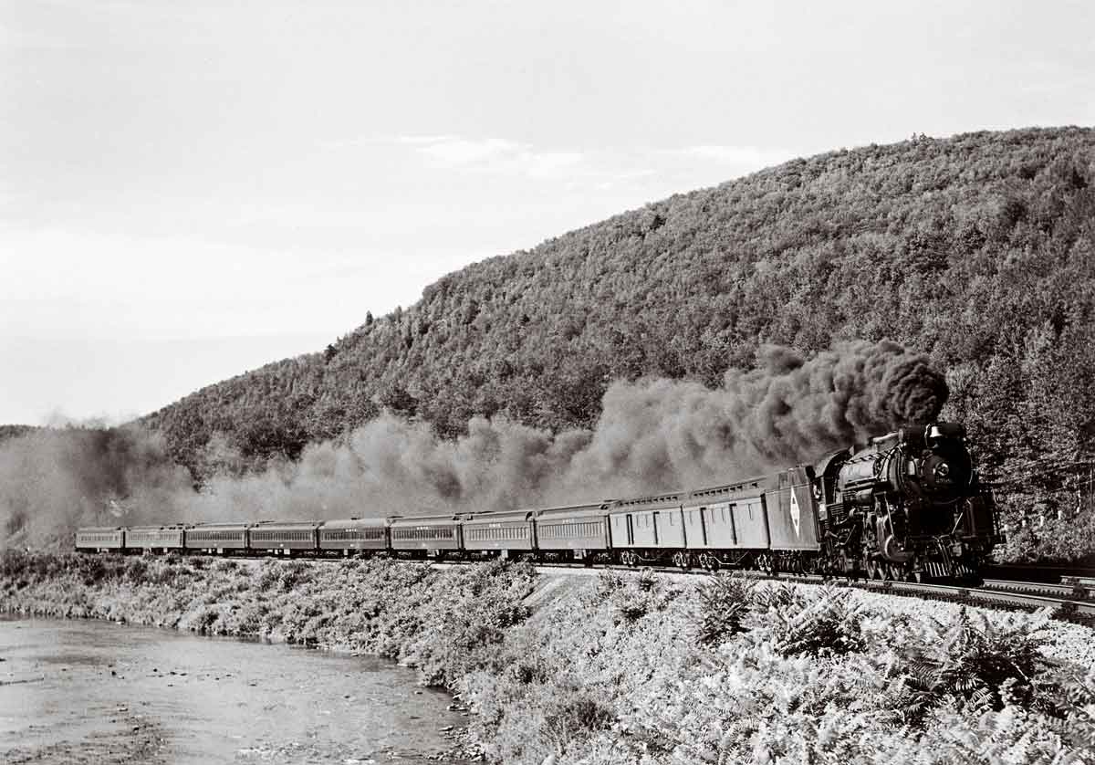 A black and white photo of a train turning a corner with black smoke coming out of its chimney