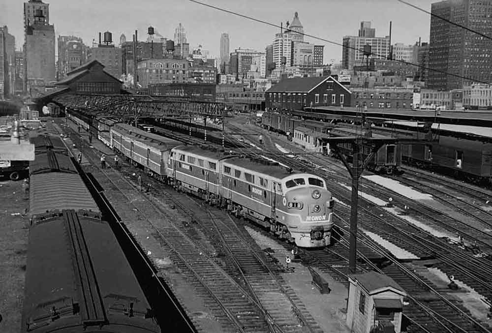 a diesel train pulling passenger cars with a train station in the background