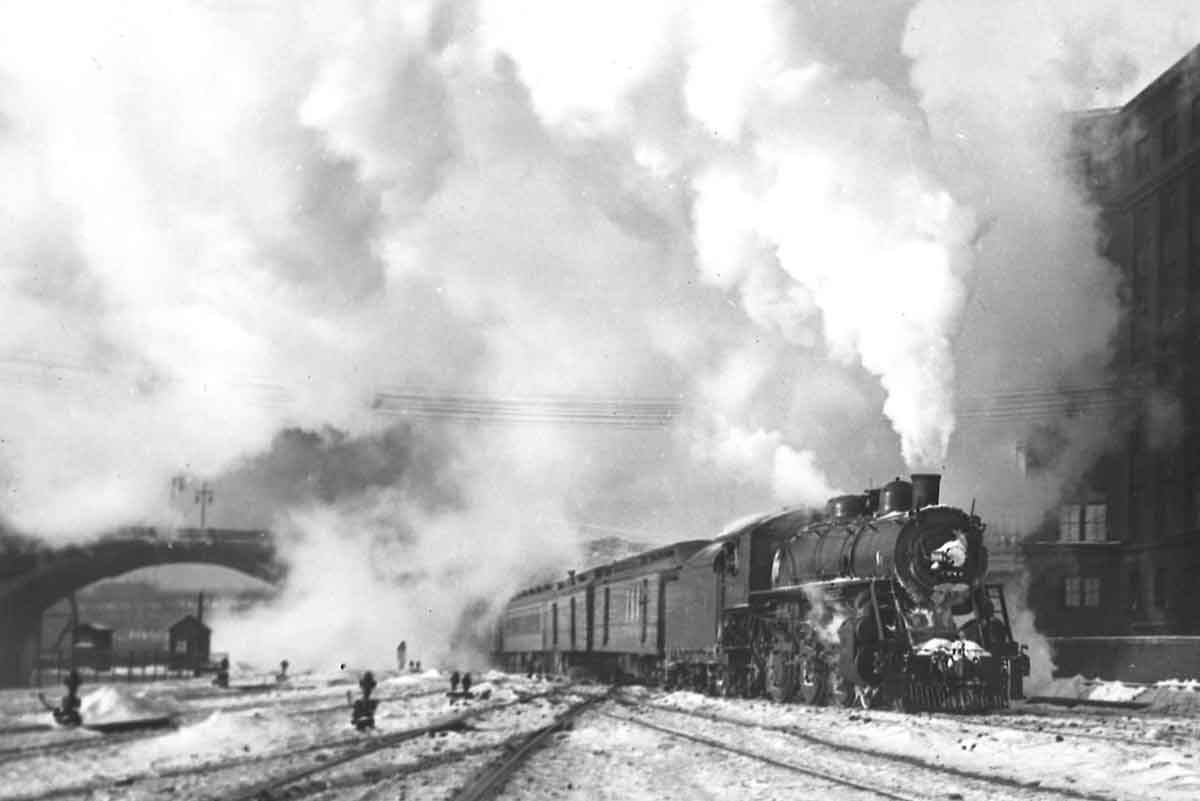 A black and white photo of a train in front of a building with big white smoke coming out of its chimney