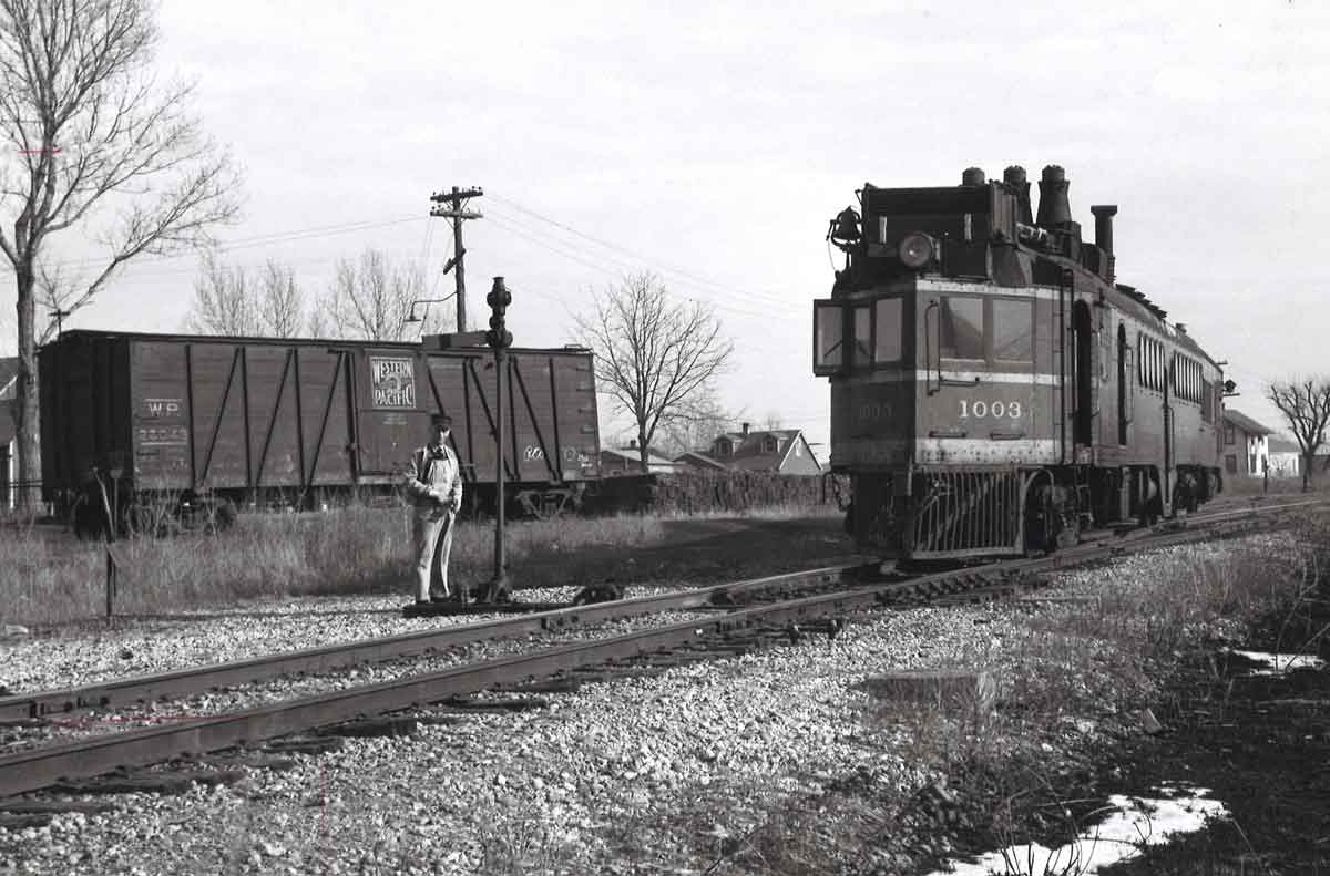 A black and white photo of a man standing by a stop light in front of a train