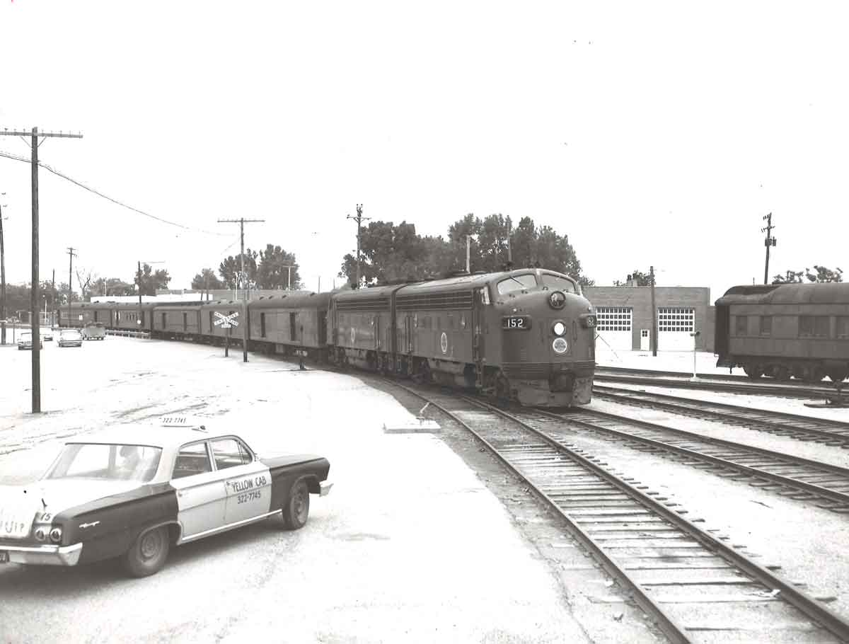 A black and white photo of a train parked on the tracks next to a taxi cab